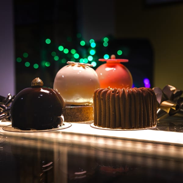 Four gourmet desserts, including chocolate and white glazed cakes, displayed on a counter with green bokeh lights.