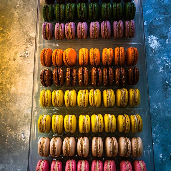 Rows of colorful macarons in green, purple, orange, brown, yellow, beige, and pink in clear trays.