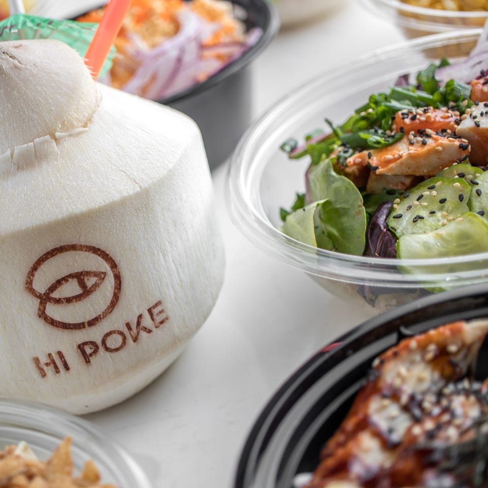 A Hi Poke branded coconut drink with a straw next to several poke bowls.